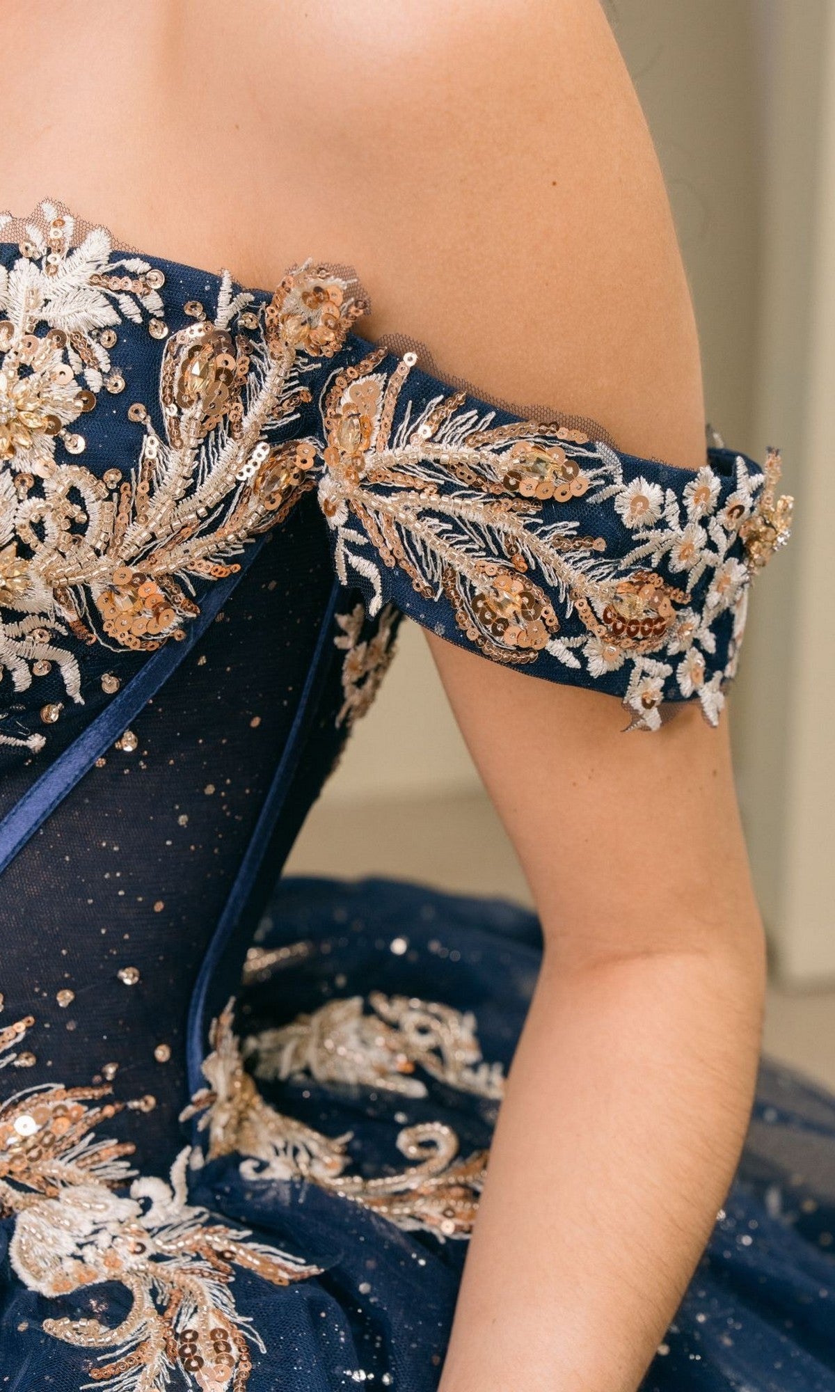 A person wearing the Quinceanera Dress 8390JQ by Cinderella Couture, an off-shoulder navy blue gown adorned with gold embroidery, sequins, and sheer fabric.